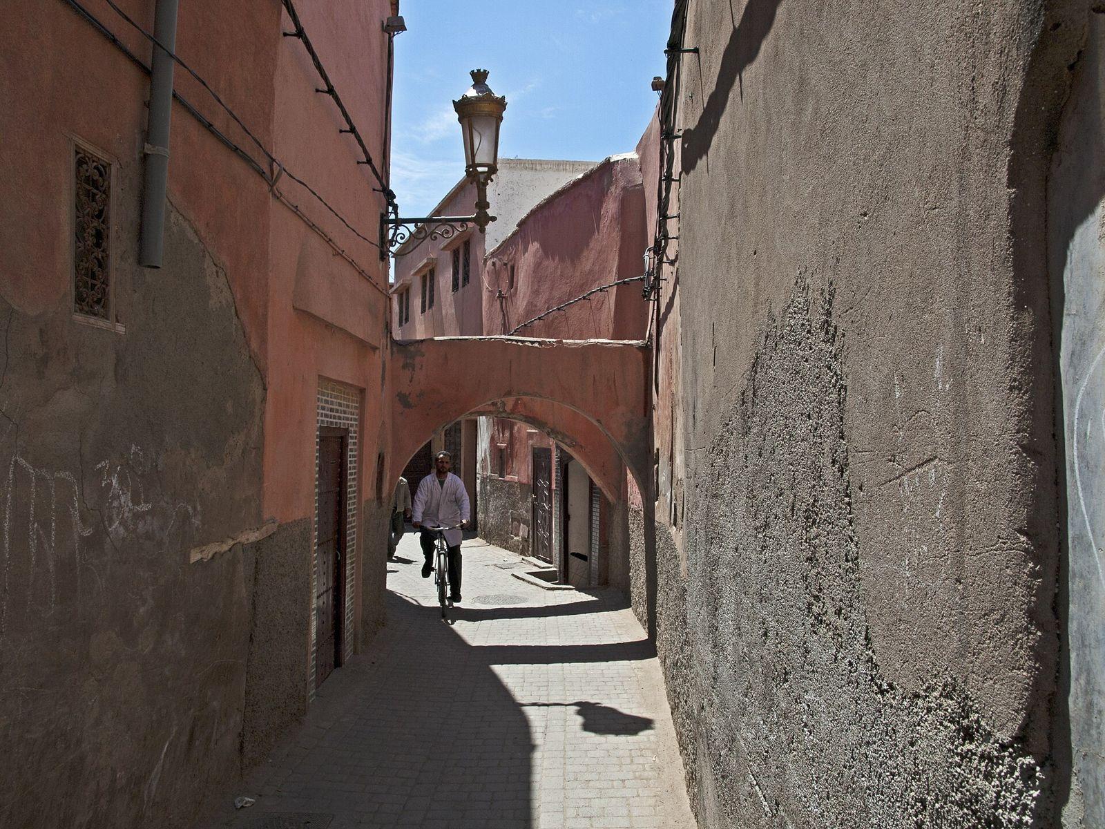Narrow medina alley in Marrakech with pink walls, an archway, a street lamp and a man on a bicycle