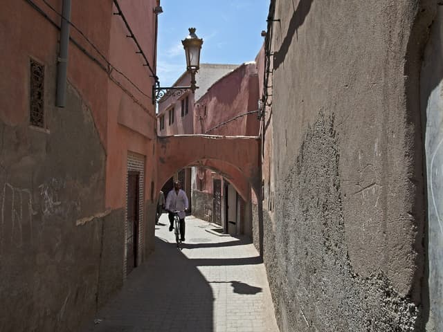 Narrow medina alley in Marrakech with pink walls, an archway, a street lamp and a man on a bicycle